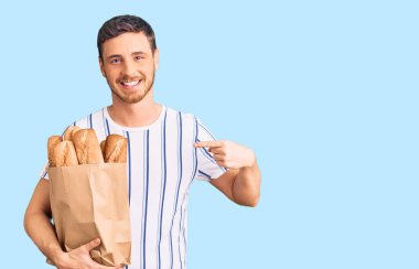 Elinde ayı olan yakışıklı genç adam, elinde kağıt torbayla elinde ekmek, gülümsüyor, mutlu bir şekilde elini ve parmağını gösteriyor. 