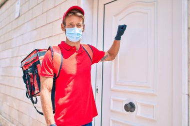 Beyaz teslimatçı kırmızı üniforma ve teslimat çantası ve koronavirüs güvenlik maskesi takıyor.