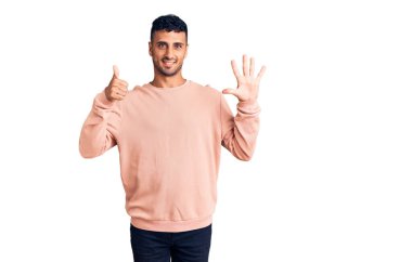 Latin asıllı genç adam gündelik giysiler giyip altı numaralı parmaklarıyla işaret ederken kendinden emin ve mutlu bir şekilde gülümsüyor.. 