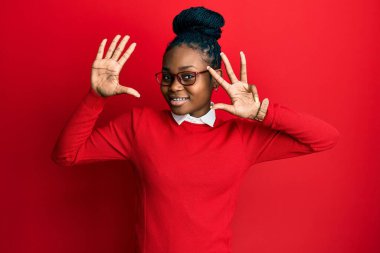 Genç Afro-Amerikalı kadın, günlük kıyafetler ve gözlük takıyor ve dokuz numaralı parmaklarıyla işaret ediyor, gülümsüyor ve mutlu.. 