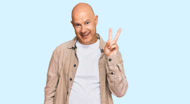 Orta yaşlı, kel bir adam, günlük kıyafetler giyip iki numaralı parmaklarıyla işaret ederken kendinden emin ve mutlu bir şekilde gülümsüyor.. 