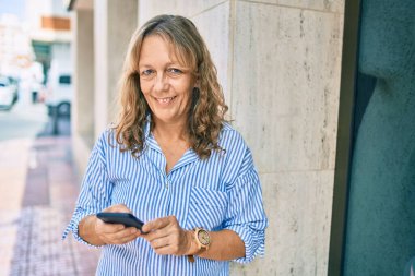 Orta yaşlı beyaz kadın şehirde akıllı telefon kullanarak mutlu bir şekilde gülümsüyor..