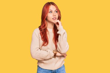 Young redhead woman wearing casual winter sweater with hand on chin thinking about question, pensive expression. smiling and thoughtful face. doubt concept. 