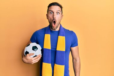 Sakallı yakışıklı adam futbol fanatiği tezahürat yapıyor topu tutuyor ve ağzı açık şaşkınlık ve şaşkınlık içinde şaşırıyor, inançsız yüz ifadesi. 