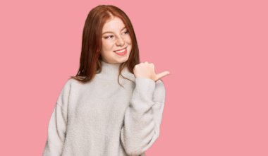 Kış süveteri giyen, neşeli yüzüyle gülümseyen ve baş parmağıyla yan tarafı gösteren genç, okumuş bir kadın.. 