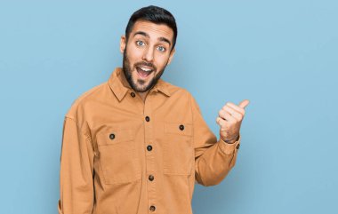 Gündelik kıyafetler giyen, gülümseyen, gülümseyen ve başparmağıyla yan tarafı gösteren İspanyol genç adam.. 