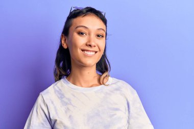 Gündelik kıyafetler giyen genç bir kadın pozitif ve mutlu görünüyor. Kendine güvenen bir gülümsemeyle ayakta duruyor ve dişlerini gösteriyor. 