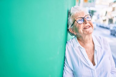 Yaşlı, yaşlı, kır saçlı, gülümseyen, açık havada duvara yaslanan yaşlı bir kadın.