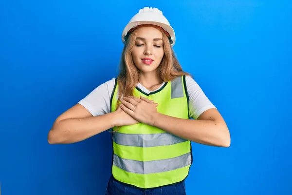 Beautiful blonde caucasian woman wearing architect hardhat smiling with hands on chest, eyes closed with grateful gesture on face. health concept.