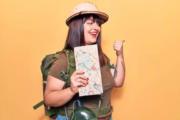 Young plus size woman wearing explorer hat holding map pointing thumb up to the side smiling happy with open mouth