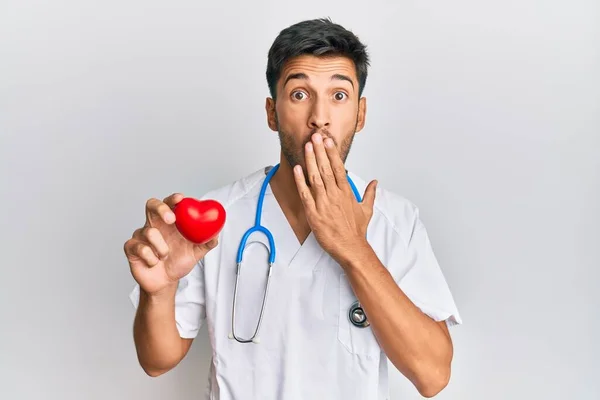 Joven Hombre Guapo Vistiendo Uniforme Médico Sosteniendo Corazón Cubriendo Boca — Foto de Stock