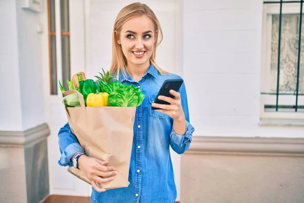 Joven Chica Rubia Sonriendo Feliz Usando Teléfono Inteligente Sosteniendo Grocries —  Fotos de Stock