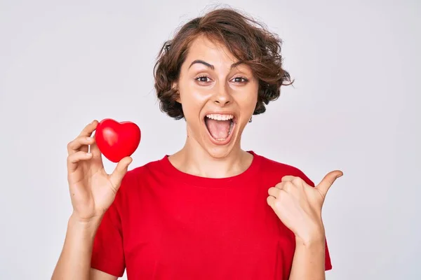 Joven Mujer Hispana Sosteniendo Corazón Señalando Pulgar Hacia Costado Sonriendo —  Fotos de Stock