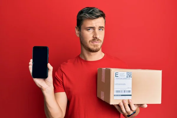 Handsome Caucasian Man Holding Delivery Package Showing Smartphone Screen Clueless — Stock Photo, Image