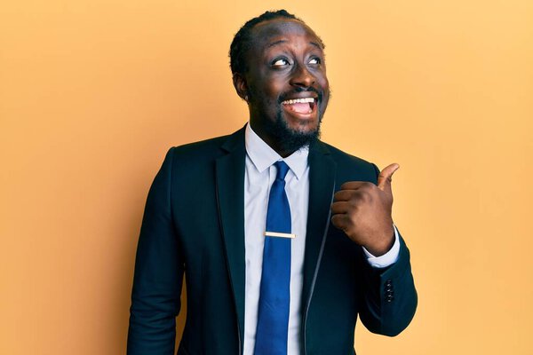 Handsome young black man wearing business suit and tie pointing thumb up to the side smiling happy with open mouth 