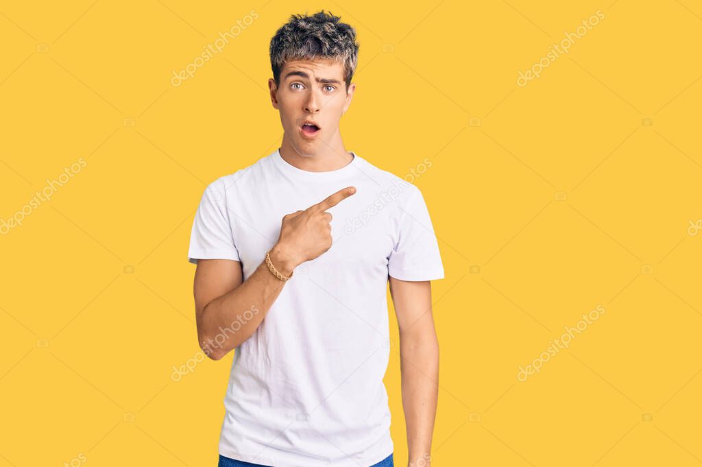 Young handsome man wearing casual white tshirt surprised pointing with finger to the side, open mouth amazed expression. 
