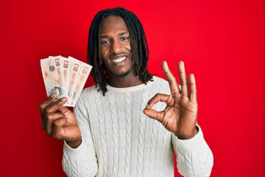 Saçları örgülü Afrika kökenli Amerikalı bir adam elinde 10 pound banknot tutuyor. Parmaklarıyla imza atıyor, gülümsüyor ve mükemmel bir sembol. 