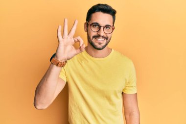 Gündelik kıyafetler ve gözlük takan genç İspanyol adam gülümsüyor. El ve parmaklarıyla 