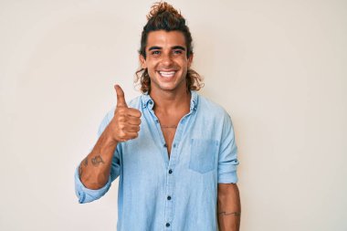 Yaz stili giyinmiş genç İspanyol adam mutlu ve pozitif gülümsüyor, başparmağıyla mükemmel ve onay işareti yapıyor. 