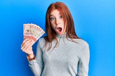 Young red head girl holding 10 colombian pesos banknotes scared and amazed with open mouth for surprise, disbelief face 