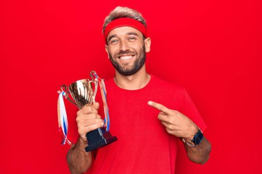 Sakallı, genç, sarışın sporcu izole edilmiş kırmızı arka planda zafer kupası kazanıyor.