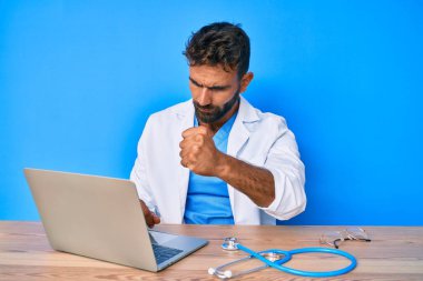 Young hispanic man wearing doctor uniform working at the clinic annoyed and frustrated shouting with anger, yelling crazy with anger and hand raised 