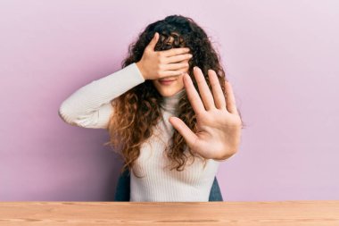 Latin kökenli genç bir kız, gündelik giysiler içinde masada oturuyor, elleriyle gözlerini kapatıyor ve üzgün ve korku dolu ifadelerle jestleri durduruyor. utangaç ve olumsuz kavram. 