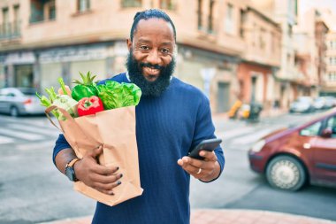 Sakallı Afrikalı Amerikalı adam elinde süpermarketten alınmış kağıt torbalarla akıllı telefon kullanıyor.
