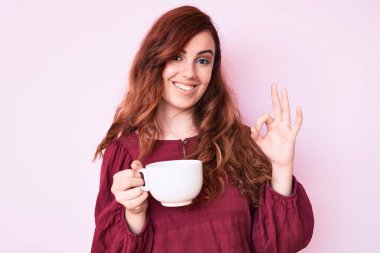 Genç, güzel bir kadın elinde kahve tutuyor. Parmaklarıyla imza atıyor. Gülümsüyor. 