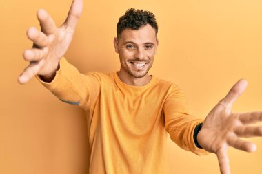 Gündelik giysiler giymiş genç İspanyol adam kameraya bakıp sarılmak için kollarını açmış gülümsüyor. Mutluluğu kucaklayan neşeli bir ifade.. 