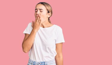 Gündelik beyaz tişört giyen genç, güzel sarışın kadın ağzıyla ağzını kapatmaktan sıkılmış. Huzursuz ve uykulu.. 