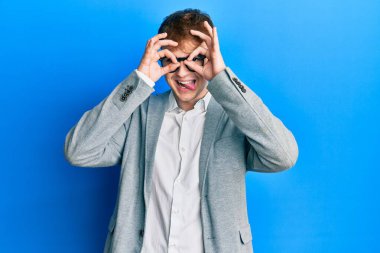 Zarif kıyafetler giyen ve gözlük takan genç beyaz bir adam dürbün gibi jest yapıyor. Dürbün gibi dilini dışarı çıkarıyor, gözler parmaklarından bakıyor. çılgın ifade. 