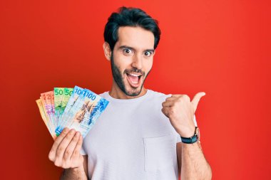 İspanyol asıllı genç adam İsviçre frangı banknotları tutuyor başparmağıyla yan tarafı işaret ediyor, ağzı açık gülümsüyor. 