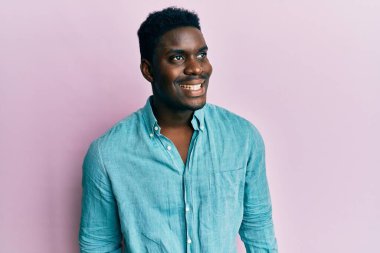 Yakışıklı, sıradan kıyafetler giyen, yüzünde gülümseme ve doğal ifadeyle yan yana bakan siyahi bir adam. Kendinden emin gülen. 