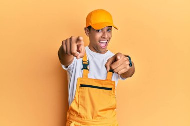 Sarı arka plan üzerine tamirci üniforması giyen yakışıklı Afro-Amerikalı genç adam seni ve kamerayı parmaklarıyla gösteriyor, gülümsüyor, pozitif ve neşeli. 