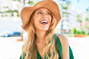 Yaz şapkası takmış, mutlu bir şekilde gülümseyen güzel sarışın bir kadın. Şehrin caddesinde yüzünde gülümsemeyle dikilirken.