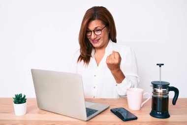 Orta yaşlı esmer İspanyol iş kadını ofiste zaferi mutlu bir gülümseme ve ellerini kaldırarak kutluyor. 