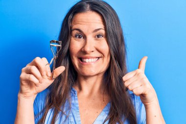 Genç, güzel esmer bir kadın mavi arka planda kirpik bigudisi tutuyor mutlu ve pozitif gülümsüyor, başparmağını kaldırıyor ve mükemmel ve onay işareti yapıyor.
