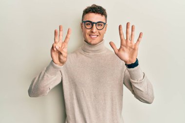 Balıkçı yaka kazak giyen İspanyol genç adam gülümseyerek kendinden emin ve mutlu bir şekilde sekiz numarayı gösteriyor ve işaret ediyor.. 