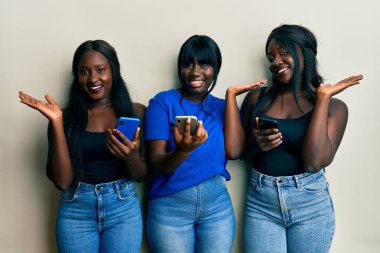 Üç Afro-Amerikalı arkadaş akıllı telefon kullanıyor neşeli bir şekilde gülümsüyor ve avuç içiyle kameraya bakıyor.. 