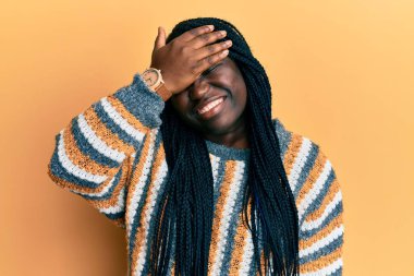 Saçları örgülü, sıradan bir süveter giyen, gülümseyen ve yüzünde şaşkınlıktan gözleri kapayan genç siyahi bir kadın. kör kavram. 