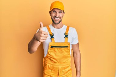 Sarı arka planda tamirci üniforması giyen yakışıklı genç adam mutlu bir el hareketi yapıyor. Kameraya bakıp başarıyı gösteren ifade. 