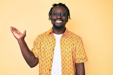 Saçları örgülü, neşeli gülümseyen, avuç içiyle kameraya bakan Afro-Amerikan genç adam.. 