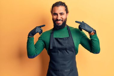 Genç Arap adam berber önlüğü takıyor. Yüzünde bir gülümsemeyle kendinden emin görünüyor. Kendini gururlu ve mutlu bir şekilde gösteriyor.. 