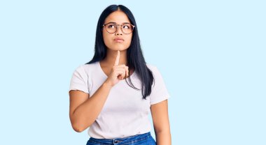 Genç, güzel Asyalı bir kız günlük kıyafetler ve gözlük takıyor şüpheye odaklanmış, çenesinde parmak olan ve kafasını kaldırıp merak eden. 