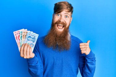 Genç İrlandalı kızıl saçlı adam Yuan Çin banknotlarını tutarken başparmağıyla yan tarafı işaret ediyor ve ağzı açık gülümsüyor. 