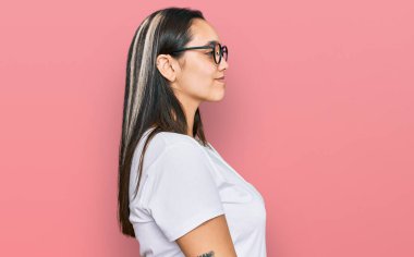 Latin kökenli genç bir kadın günlük beyaz tişört giyiyor. Yan tarafa bakıyor, rahat bir görünüm takınıyor ve kendine güvenen bir gülümsemeyle poz veriyor.. 