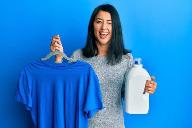 Güzel Asyalı genç kadın elinde mavi tişört ve deterjan şişesiyle kameraya bakıyor. Seksi bir ifadeyle, neşeli ve mutlu bir yüzle.. 