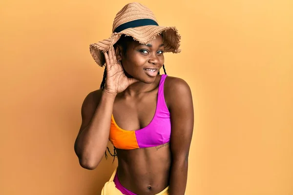 Young african american woman wearing bikini and summer hat smiling with hand over ear listening and hearing to rumor or gossip. deafness concept.