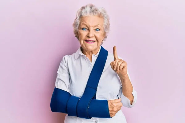 Senior grey-haired woman wearing arm on sling smiling with an idea or question pointing finger with happy face, number one